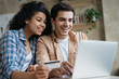 Portrait of beautiful multiracial couple shopping online, using laptop computer.  Happy friends ordering food on website. Smiling African America woman holding credit card, looking at digital screen 