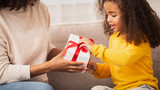 Fototapeta Panele - Little Girl Receiving Gift From Mother Sitting On Couch Indoor