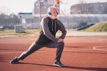 An Older Man Stretching His Legs At The Basketball Court Wearing Headphones.sportsperson