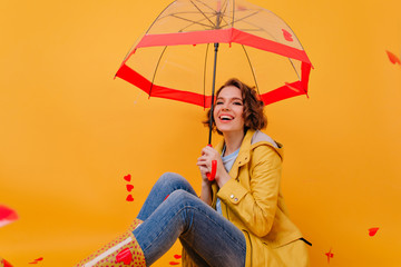 Magnificent young woman in jeans and yellow coat posing under parasol. Indoor photo of fashionable white girl enjoying photoshoot in autumn day.