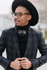 Wall Mural - Pensive male model with dark skin looking away during outdoor photoshoot. Gorgeous african young man in vintage hat posing on blur town background.