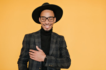 Happy black young man wears elegant dark suit posing with pleased smile. Indoor photo of relaxed male model in glasses enjoying photoshoot.