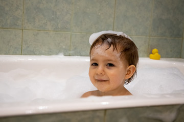 Wall Mural - Cute little toddler girl smiles taking a bath with a soap foam arround and her head. close up, soft focus, backgound is green bathroom in blur