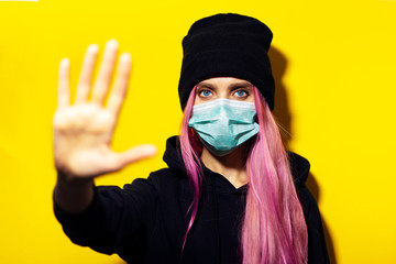 Wall Mural - Studio portrait of young girl with pink hair and blue eyes, wearing medical flu mask, hooded sweater and beanie hat, showing Stop gesture on yellow background.