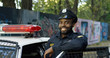 © VAKSMANV - Portrait of handsome African American young policeman in uniform and cap. Front view of police officer smiling at camera outdoors. Cop on the background with police car and old lane.