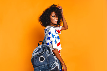 Wall Mural - Happy young woman with national flag of USA. American dream. Hilariously beautiful girl with curly hair in denim jeans, backpack and white t-shirt at yellow background. 