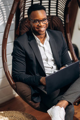 Wall Mural - Stylishly dressed African American businessman is sitting in a chair in the creative office with a laptop in his hands