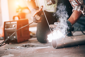 Welder in protective uniform and mask welding metal pipe on the industrial table while sparks flying.