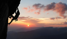 Male Climber In Rock Climbing Equipment, Pulling Up And Doing Next Step On High Cliff Reaching Rocky Top. Side View. Panoramic View Of Mountains And Amazing Pink Sky With Sunset And Clouds. Copy Space