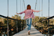 © Aleksandra - slender young girl in a red shirt and jeans with her arms outstretched is walking on a suspended cable-stayed old wooden bridge, traveling alone and in pleasure