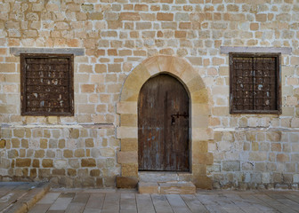 Old weathered arched wooden door and two closed rusted wrought iron windows on bricks stone wall and tiled stone floor