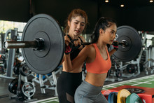 Asian Personal Trainer Coaching A Bodybuilding Woman To Perform The Exercise Squat With Barbell In The Fitness Gym. Weightlifting And Workout Training Concept