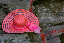 A Bright And Rich Pink Hat With A Stripe Pattern And A Matching Satin Ribbon Bow Hangs On Large Stone Rocks In A Darkened Place With Sunlight.