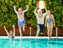 Funny View Of Couples Of Senior People Playing In The Blue And Transparent Water Of The Swimming Pool. Man Jumps Into The Pool With A Large Inflatable Ball