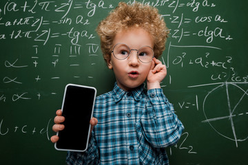 Wall Mural - Smart child holding smartphone with blank screen and touching glasses near chalkboard with mathematical formulas