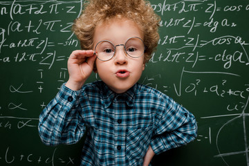 smart kid touching glasses and standing with hand on hip near chalkboard