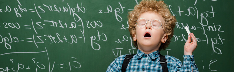 Wall Mural - panoramic shot of curly kid in glasses sneezing near chalkboard with mathematical formulas