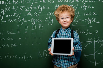 Wall Mural - happy and smart kid in glasses holding digital tablet with blank screen near chalkboard with mathematical formulas