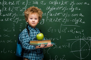Wall Mural - curly kid holding books and fresh apple near chalkboard with mathematical formulas