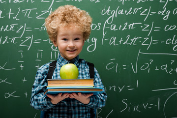Wall Mural - curly kid holding books and apple near chalkboard with mathematical formulas