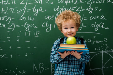 Wall Mural - cheerful kid holding books and apple near chalkboard with mathematical formulas