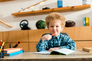 Wall Mural - happy and curly kid holding pen near notebook and books on desk