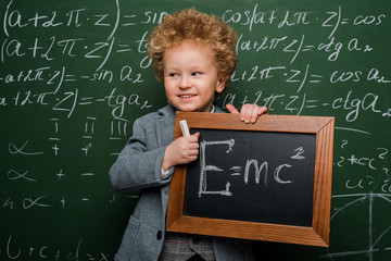 Wall Mural - cheerful kid in suit and bow tie holding small blackboard with formula near chalkboard