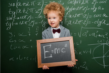 Wall Mural - smart child in suit and bow tie holding small blackboard with formula near chalkboard