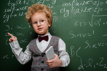 Wall Mural - smart child in suit and bow tie holding smartphone and pointing with finger near chalkboard with mathematical formulas