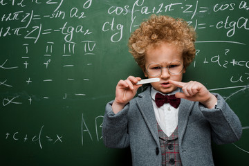 Wall Mural - smart child in suit with bow tie pointing with finger at chalk near chalkboard with mathematical formulas
