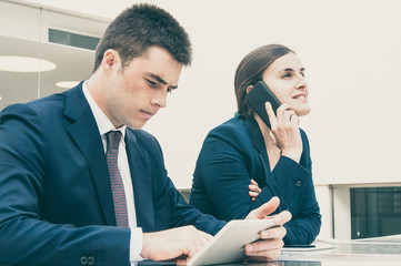 Colleagues using tablet and calling on phone at desk outdoors. Business man and woman wearing formal clothes and sitting at cafe table. Business and communication concept.