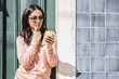 © Mangostar - Pensive woman using smartphone outdoors. Pretty young lady standing at door and wall. Communication concept. Front view.