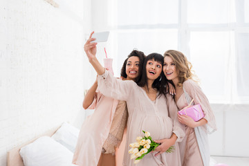 Wall Mural - multiethnic girlfriends and happy pregnant woman holding milkshake, flowers and gift while taking selfie on baby shower