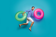 Full Length Photo Of Funky Crazy Guy Tourist Jump High Running Swimming Hold Two Colorful Lifebuoys Circles Wear Striped Sailor Shirt Shorts Flip Flops Isolated Blue Color Background