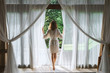A woman in a bathrobe opens the curtains in deluxe Bali hotel room overlooking the terrace and tropical trees.Woman is awoke and standing before window. Girl is opening curtains and meeting sunrise