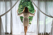 A woman in a bathrobe opens the curtains in deluxe Bali hotel room overlooking the terrace and tropical trees.Woman is awoke and standing before window. Girl is opening curtains and meeting sunrise
