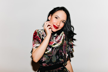 Wall Mural - Glamorous black-haired woman with bright makeup posing with surprised smile. Glad female model with dark hair touching her face and laughing on white background.