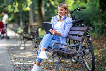 Wall Mural - Blonde woman sitting on bench with tablet on her hands and headphones over her neck