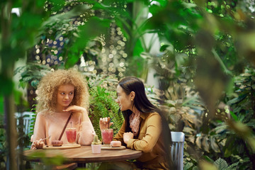 Two young women drinking juice eating dessert and talking to each other during their meeting in cafe