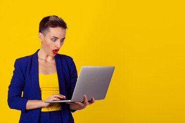 Surprised Shocked young business woman short hair using laptop looking at computer screen blown away in stupor on yellow background. Human face expression, emotion, feeling, body language, reaction