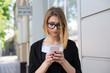 © hbrh - Bad news by sms. Portrait of annoyed stressed office young woman holding cellphone in hands on a city street looking angry by what she saw online. Mixed race model in black business attire and glasses
