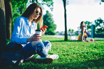 Young  millennial woman in stylish glasses watching video on smartphone and using accessory sitting near trees in park, female blogger recreating outdoors publication text via application and 4G