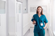 © Monkey Business - Portrait Of Smiling Female Doctor Wearing Scrubs Standing In Hospital Corridor Holding Clipboard