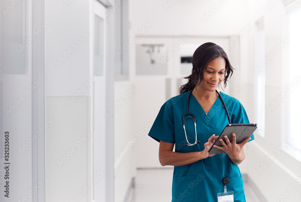 Female Doctor Wearing Scrubs In Hospital Corridor Using Digital Tablet - obrazy, fototapety, plakaty 