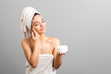 beauty woman in towel holding white jar with cream and caress her skin. grey background