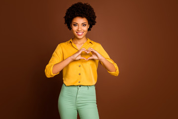 Poster - Photo of pretty dark skin wavy lady holding arms fingers in heart shape symbolizing cardiac healthy healthcare concept wear yellow shirt green trousers isolated brown color background