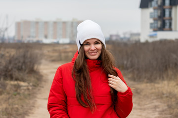 Smiling beautiful young woman with long hair in a white knitted hat and a red jacket with a hood on the street on a cloudy day against a blurred background. Girl without makeup, natural beauty