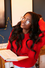 Young beautiful african american woman in a red knitted jumper holds a book and and straightens luxurious dark hair. Happy girl in christmas vacation.