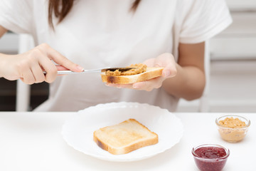 A young woman spreads peanut butter paste on bread, prepares an American sandwich.