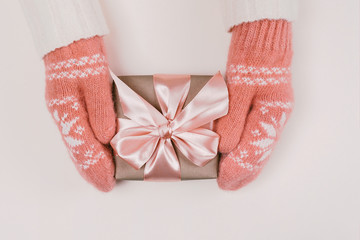 Wall Mural - Woman's hands in warm knitted mittens hold gift box wrapped with craft paper and pink ribbon on white background.